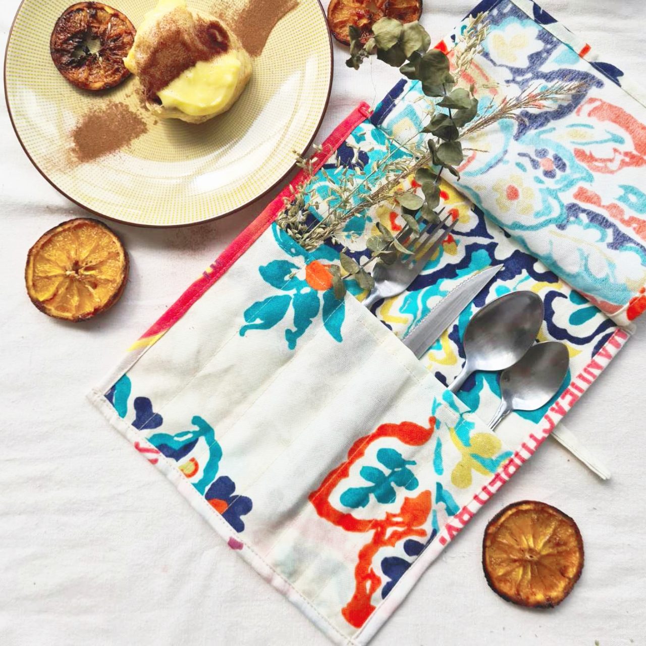 A floral katsa cutlery pocket holding a spoon and fork, on a table with cake and dried orange slices